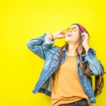 Smiling woman in sunglasses stands against vibrant yellow wall, radiating happiness.